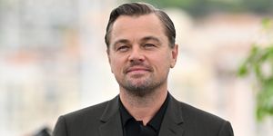 leonardo dicaprio stands and looks at the camera with a slight smile, he wears a gray suit jacket and black collared shirt, the outdoor background is blurred behind him