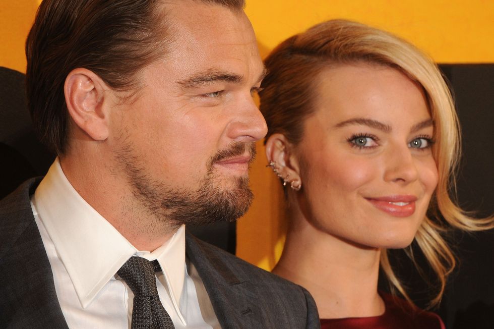 margot robbie smiles at the camera, she wears a red dress and several earrings, leonardo dicaprio is to her left and stares straight ahead in profile