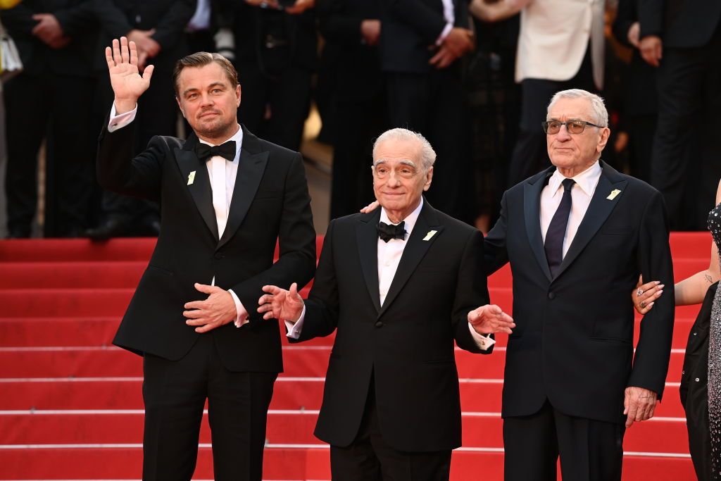 leonardo dicaprio, martin scorsese, and robert de niro stand on a red carpet, all three men are wearing black suits with white shirts and black ties, dicaprio is waving with one hand, scorsese has both hands slightly extended in front of him, and de niro has one hand on scorseses shoulder