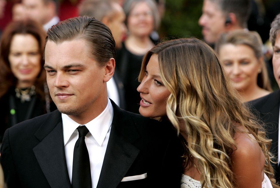 leonardo dicaprio, wearing a black suit and tie, and gisele bundchen, wearing a white dress, stand among a crowd and look off camera