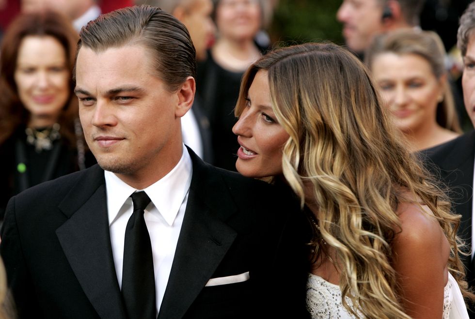 leonardo dicaprio, wearing a black suit and tie, and gisele bundchen, wearing a white dress, stand among a crowd and look off camera