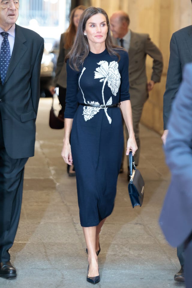 A woman in a dark dress with floral design walks through a corridor