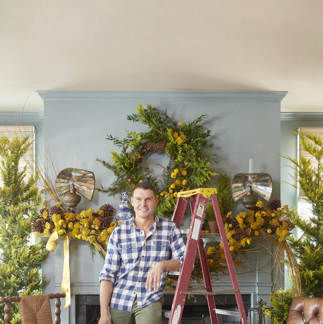 lewis miller stands in front of his mantel leaning against a ladder while he decorates for christmas