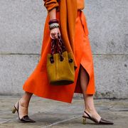 a woman wears an orange sweater, skirt, bangle bracelets, and slingback heels to fashion week while carrying a bucket bag as photographed by tyler joe