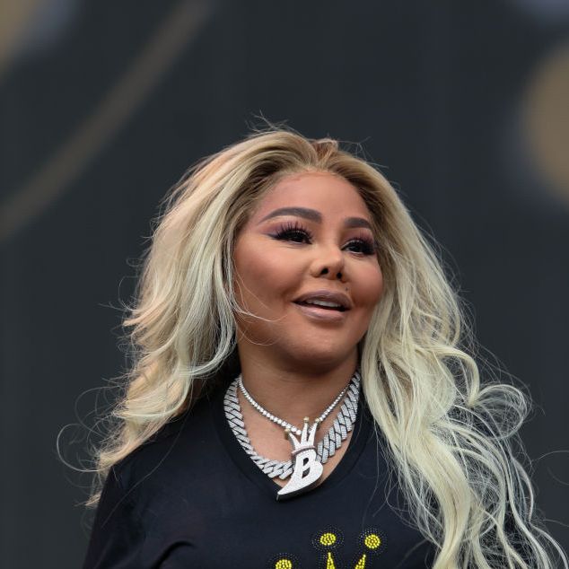 lil kim smiles while on stage, she wears a black shirt with gold rhinestones in a pattern and a large necklace with a b pendant