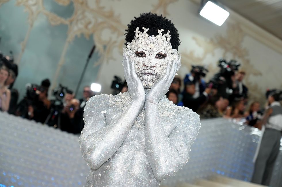 lil nas x cover in silver body paint and crystals and pearls, holding his hands to his face as he looks directly at the camera
