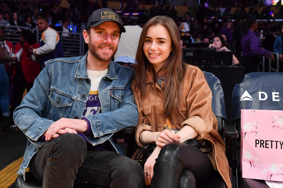 charlie mcdowell and lily collins smile at the camera while seated, they wear jackets and pants, he wears a hat