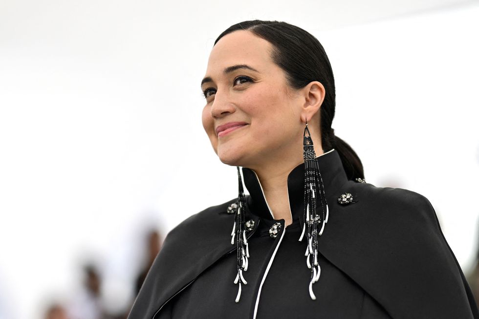a closeup of lily gladstone posing for photographers at cannes film festival, she's wearing her hair in a ponytail and is rocking a black cape dress adorned with rhinestone buttons and accessorised with black chandelier earrings