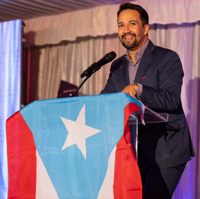 lin manuel miranda smiles and stands behind a podium draped with a puerto rican flag, he wears a dark suit with a purple unbuttoned collared shirt
