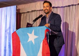 lin manuel miranda smiles and stands behind a podium draped with a puerto rican flag, he wears a dark suit with a purple unbuttoned collared shirt