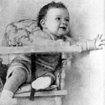 a baby sits in a high chair and touches a wall on the right as it smiles and looks up to the right