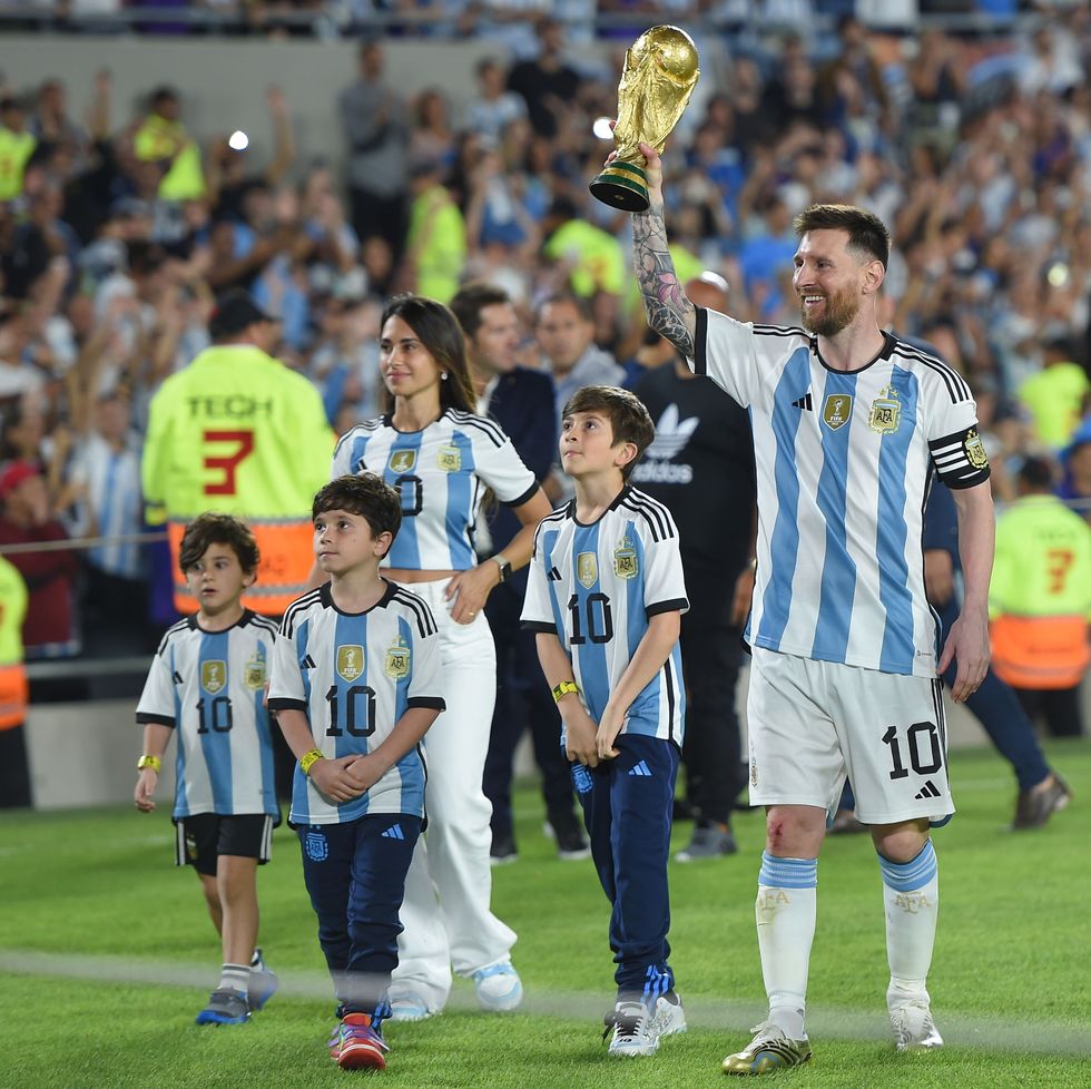 lionel messi, his wife, and three youth sons walk on a soccer field, all are wearing argentina national team jerseys and messi is holding up the fifa world cup trophy, behind them is a crowd in the stands