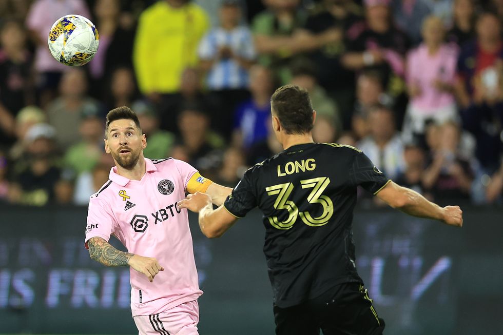 lionel messi looks at a midair soccer ball as an opponent faces him, messi wears a pink and black uniform, the opponent wears a black and gold uniform