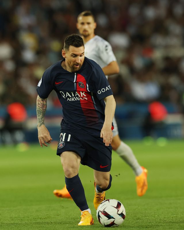 lionel messi dribbles and looks down at a soccer ball on a grass field, he wears a dark navy soccer uniform, and an opponent player watches from behind