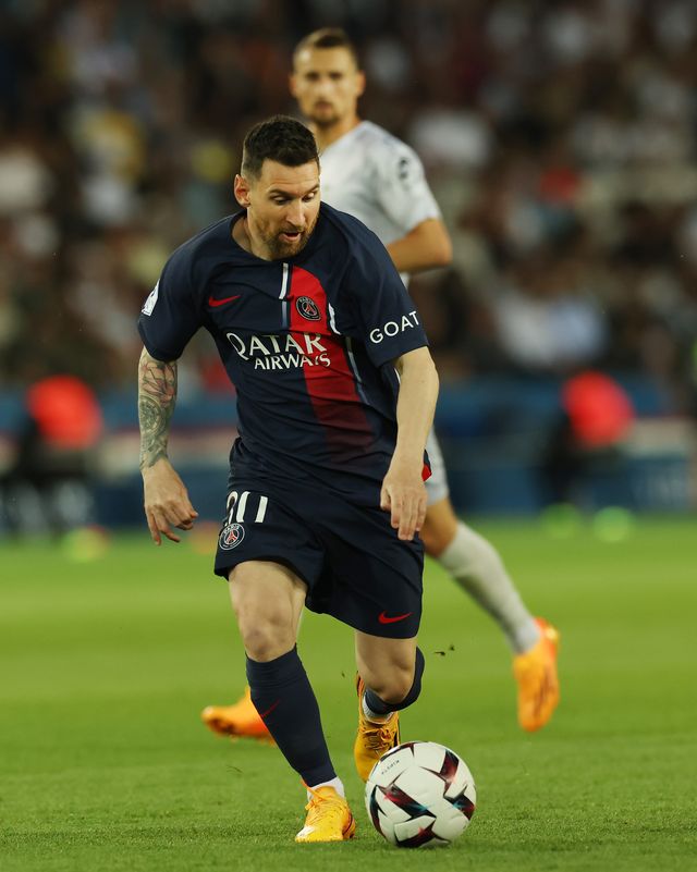 lionel messi dribbles and looks down at a soccer ball on a grass field, he wears a dark navy soccer uniform, and an opponent player watches from behind