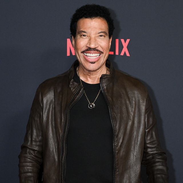 lionel richie smiles at the camera, he wears a dark brown jacket over a black shirt and necklace