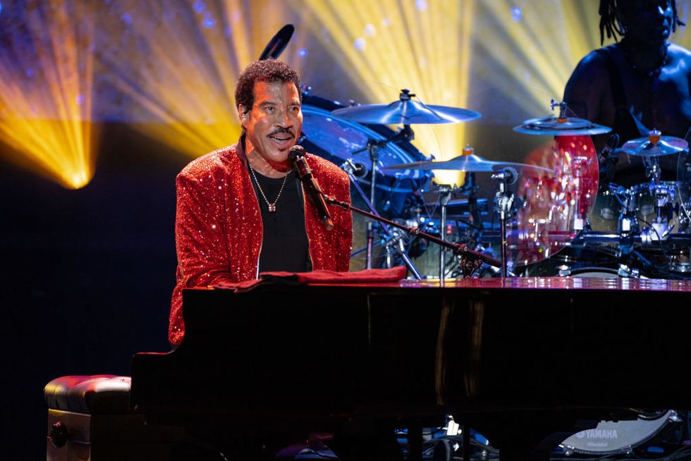 lionel richie sits behind a piano and sings into a microphone, he wears a red sparkly jacket, black shirt and silver necklace, behind him to the right is a drummer at a drumset
