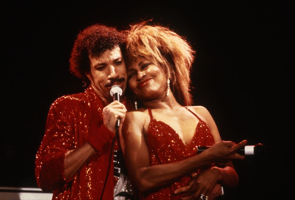 lionel richie and tina turner perform together, richie sings into a microphone he holds as turner smiles next to him