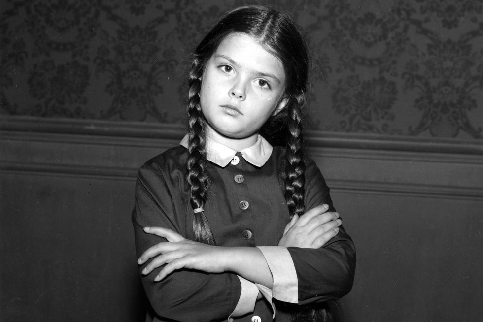lisa loring as wednesday addams, a young girl stands with arms crossed