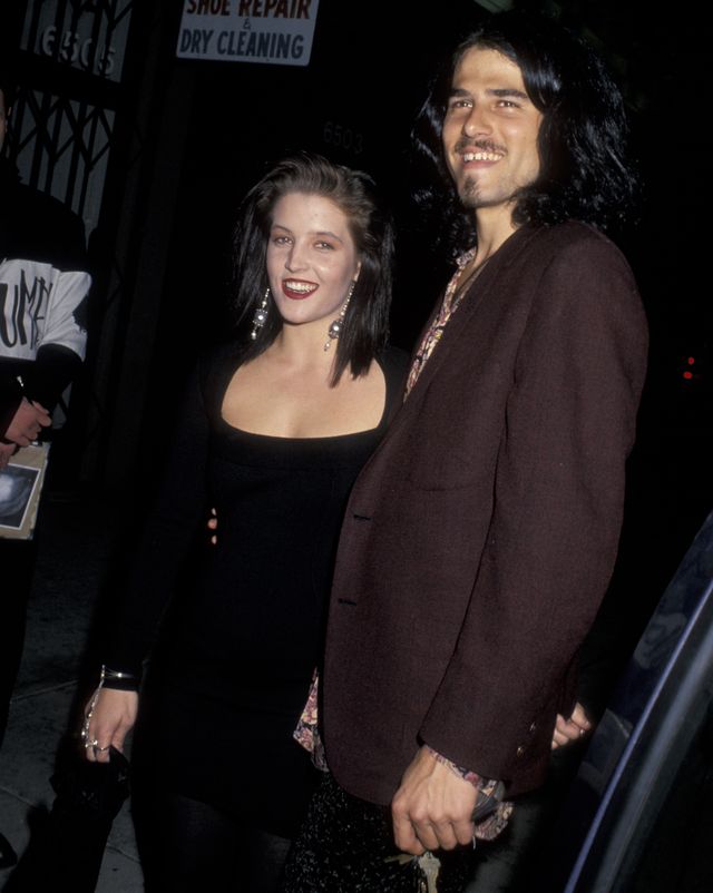 lisa marie presley and danny keough stand next to each other and smile, she wears a black scoop neck dress with long sleeves, he wears a brown suit jacket and holds a set of keys