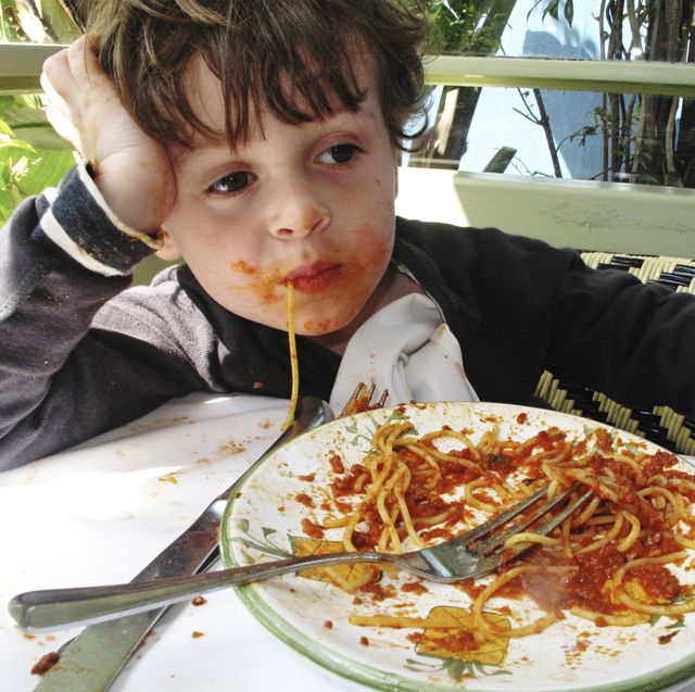 little 3 4 year old boy siting at a table eating pasta with the plate in front of him he has tomato sauce all over his face and hands and has a piece of spaghetti hanging out of his mouth