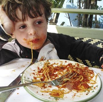 little 3 4 year old boy siting at a table eating pasta with the plate in front of him he has tomato sauce all over his face and hands and has a piece of spaghetti hanging out of his mouth