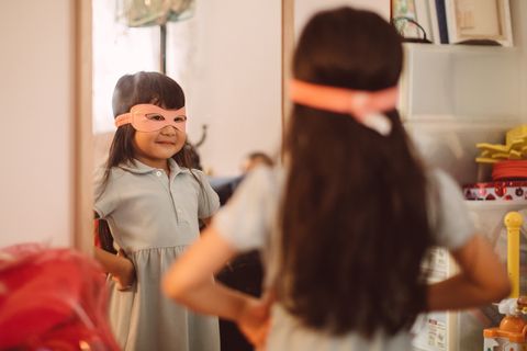 little girl wearing a superwomen mask looking into the mirror with pride