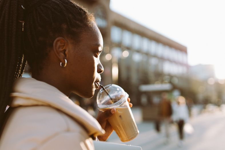 side portrait of young black woman with black braided hair in ponytail and metal earring drinking coffee from plastic cup on a spring evening