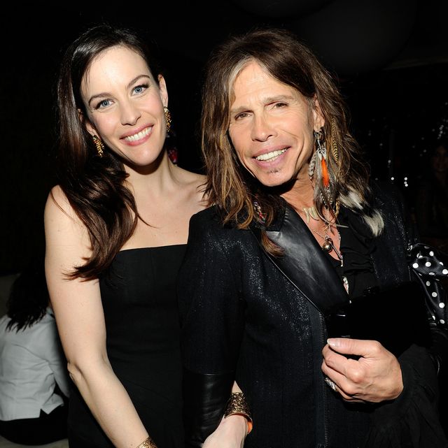 liv tyler and steven tyler stand next to each other and smile at the camera, liv is wearing a black strapless dress, steven is wearing a black suit jacket with leather lapels and ornate jewelry