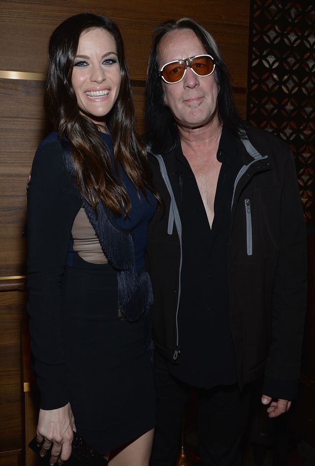liv tyler and todd rundgren stand next to each other and smile at the camera, liv is wearing a dark long sleeve dress with with a scarf, todd is wearing a black outfit with a jacket that has gray zippers and orange tinted glasses