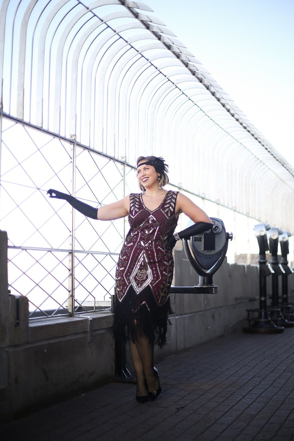 woman standing on skyscraper with mesh gate and viewfinder she is wearing 1920s attire with a black, beaded dress, long black gloves and a feathered black headband
