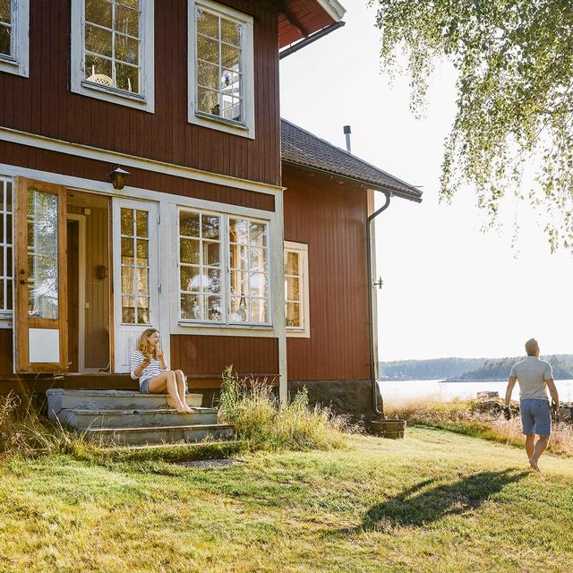 woman sitting on steps at entrance of log cabin while man walking towards lake on sunny day