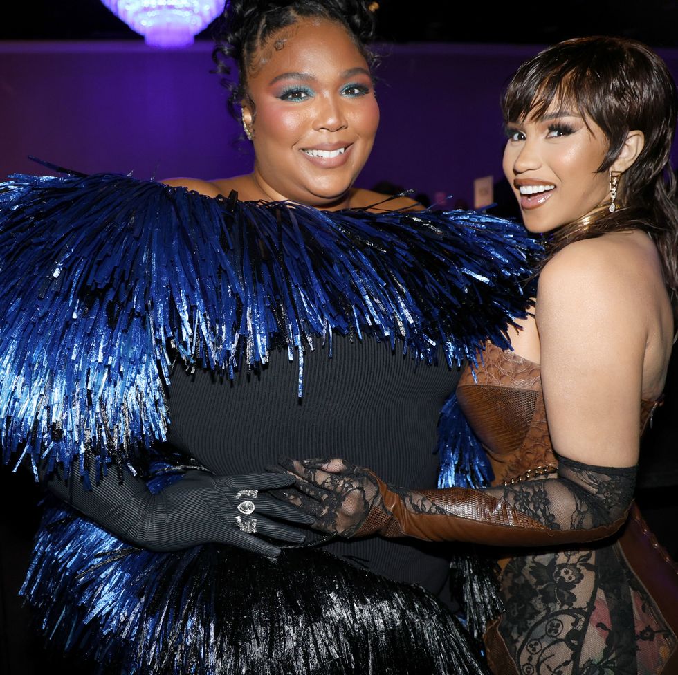 lizzo, wearing a black dress with blue grills and black gloves, holds hands and poses with cardi b, who wears a gold corset and black see through tights