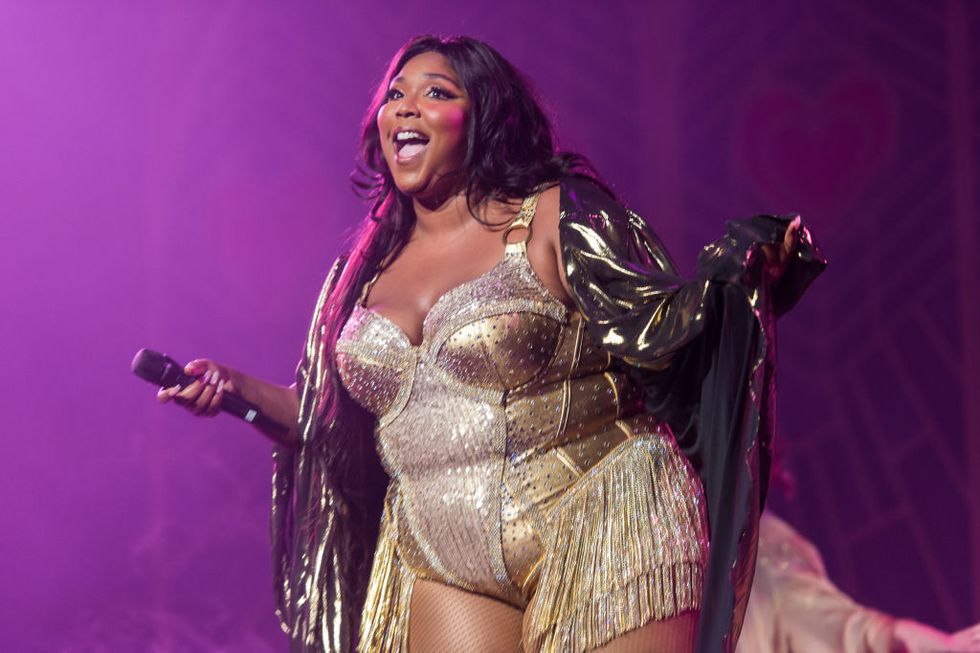 lizzo wearing a white and gold dress, holding a microphone and shouting to the audience from a stage, with purple lights in the background