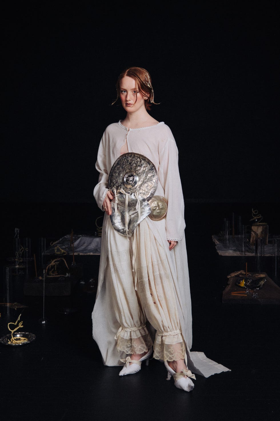Individuo con un traje histórico o teatral y un escudo de pie en un espacio poco iluminado.