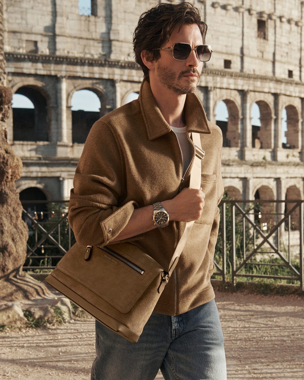 a male figure stands near the colosseum wearing a brown jacket and holding a brown bag