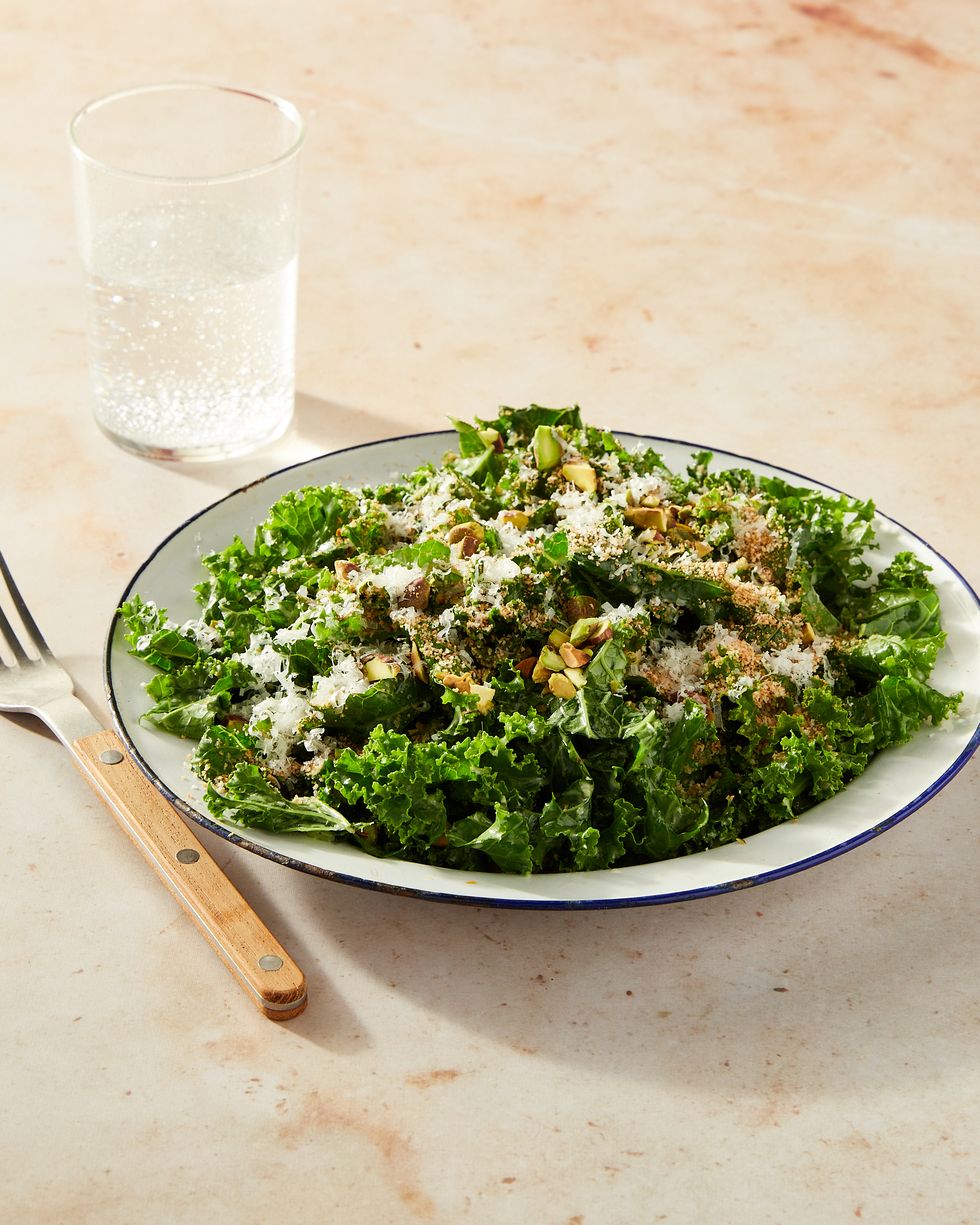 kale salad topped with cheese, breadcrumbs, and pistachios in a white bowl with a blue rim on a marble surface