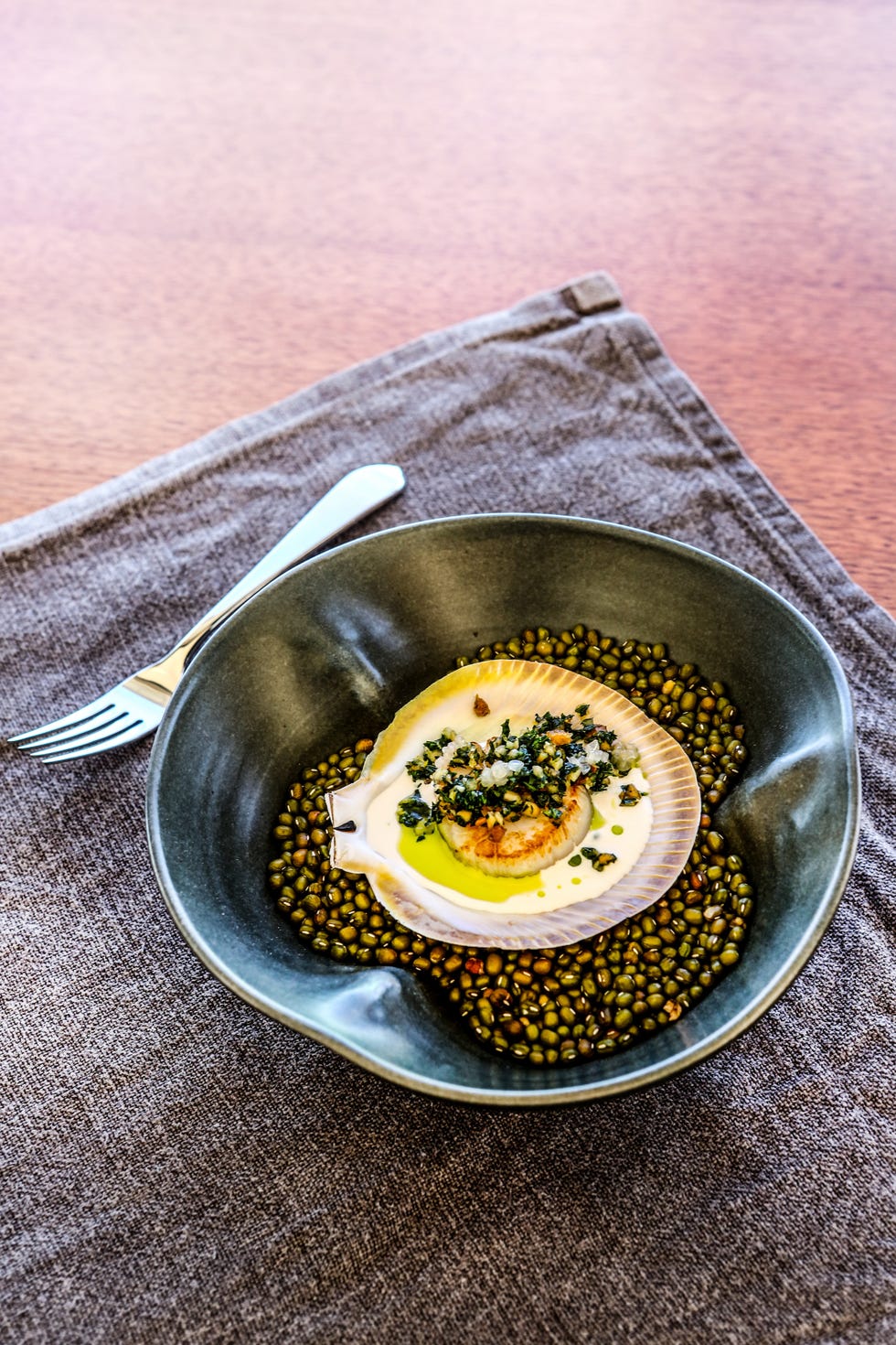 a dish featuring a scallop on a bed of green lentils garnished with herbs and olive oil