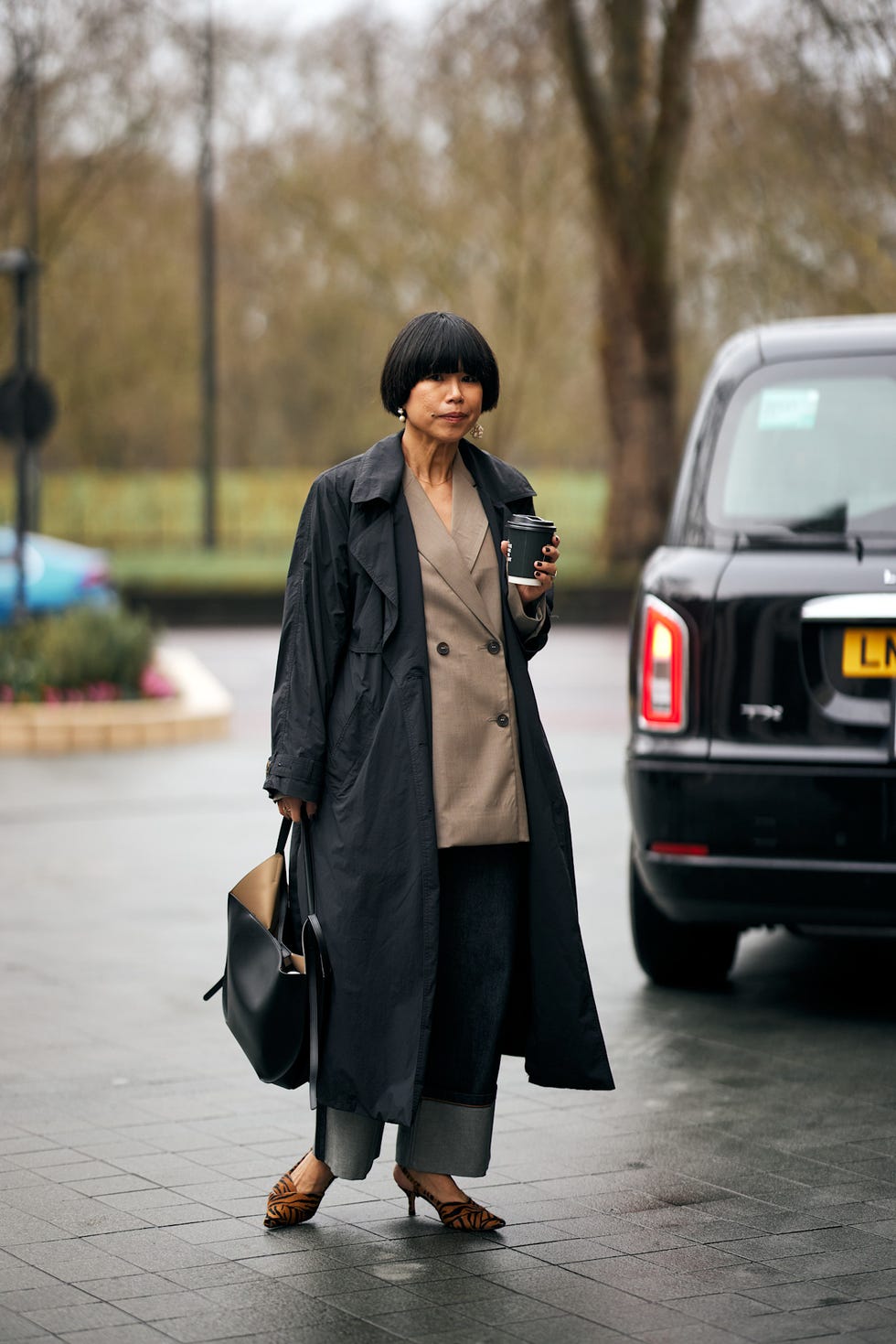 a stylish individual holding a coffee cup while standing next to a black vehicle best fringe haircuts london fashion week street style