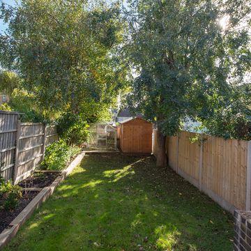 a general view of a small narrow back garden with grass lawn, railway sleeper flower beds, timber fence, green house, shed and a large tree on a sunny day