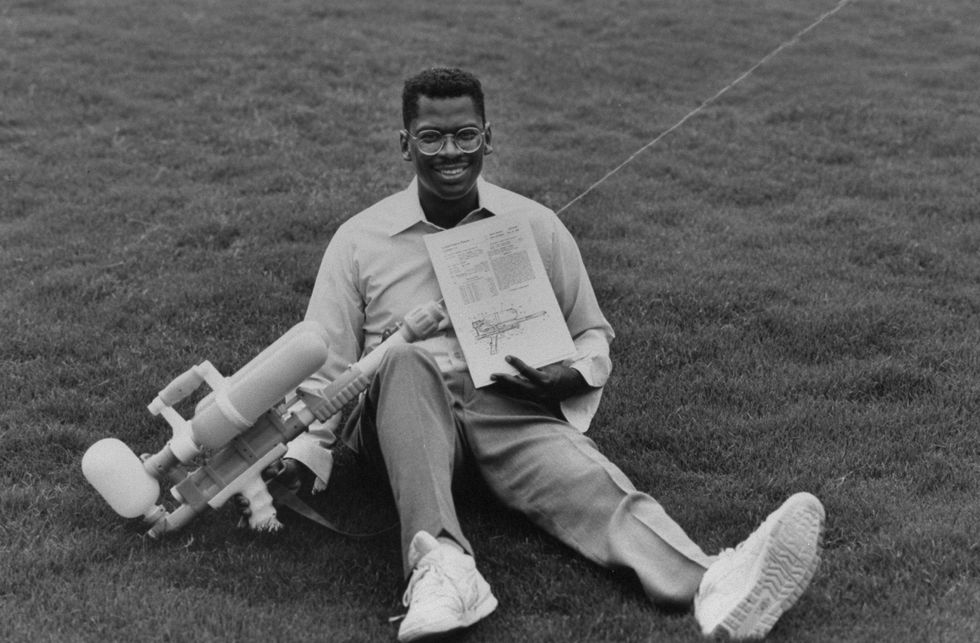 lonnie johnson sitting on a grass field with a water gun resting against his leg and a piece of paper in his hands