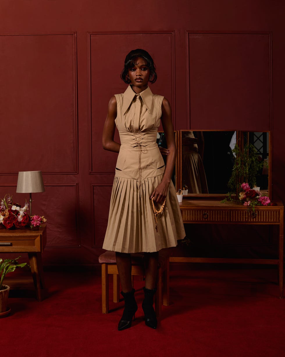 a person stands in a styled interior wearing a beige dress with pleats