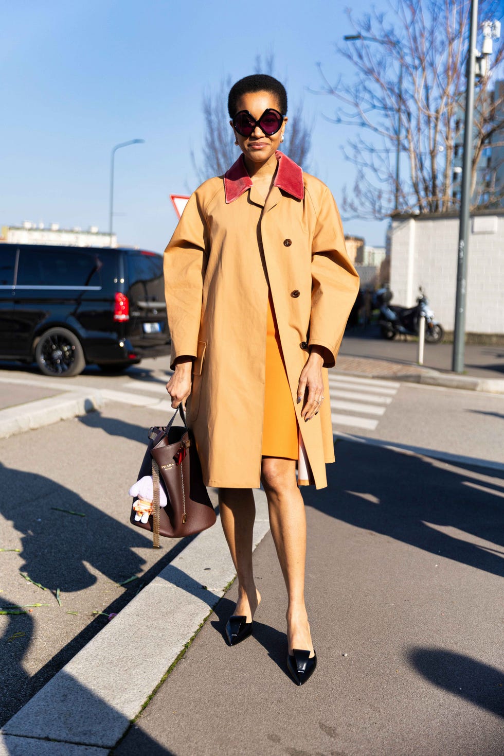 Individual dressed in an oversized coat and orange dress walking along a street with vehicles in the background