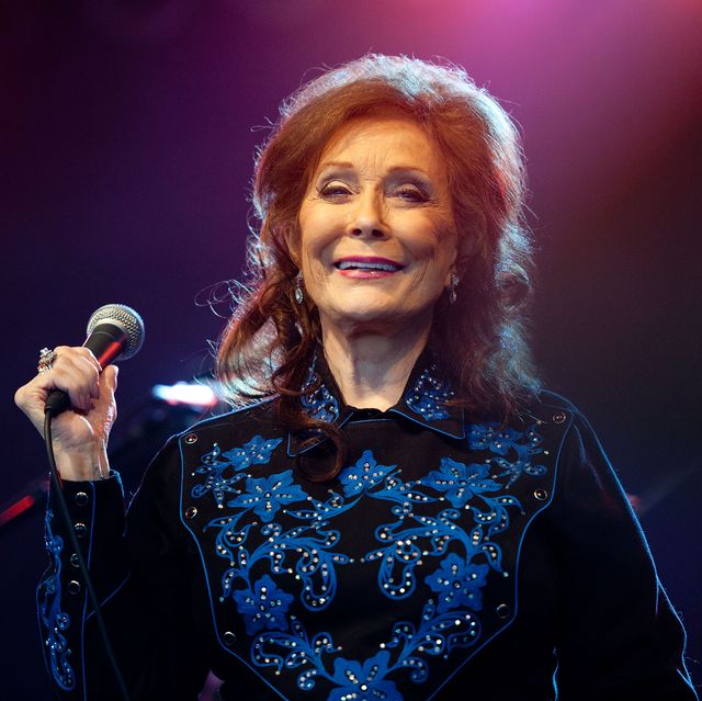 loretta lynn smiles and holds a microphone in one hand, she wears a black western shirt with a blue pattern