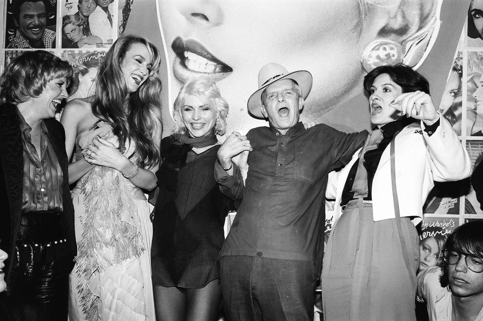 lorna luft, jerry hall, debbie harry, truman capote, and paloma picasso stand in front of a wall of posters smiling and chatting