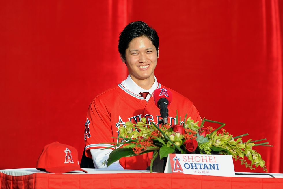 a man smiles as he sits at a table with a microphone, flowers, name card and red baseball hat, he wears a red baseball uniform for the los angeles angels over a collared shirt and red tie