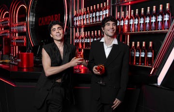 two individuals at a stylish bar holding cocktails with a camparithemed backdrop