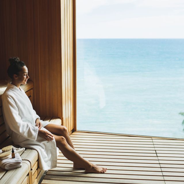young beautiful slim woman in wooden dry sauna in spa centr in white bathrobe relaxation wellness and beauty concept finnish sauna for strengthening the bodys immune system, removing toxins, resting body and mind wooden eco sauna with a large window overlooking the ocean or sea
