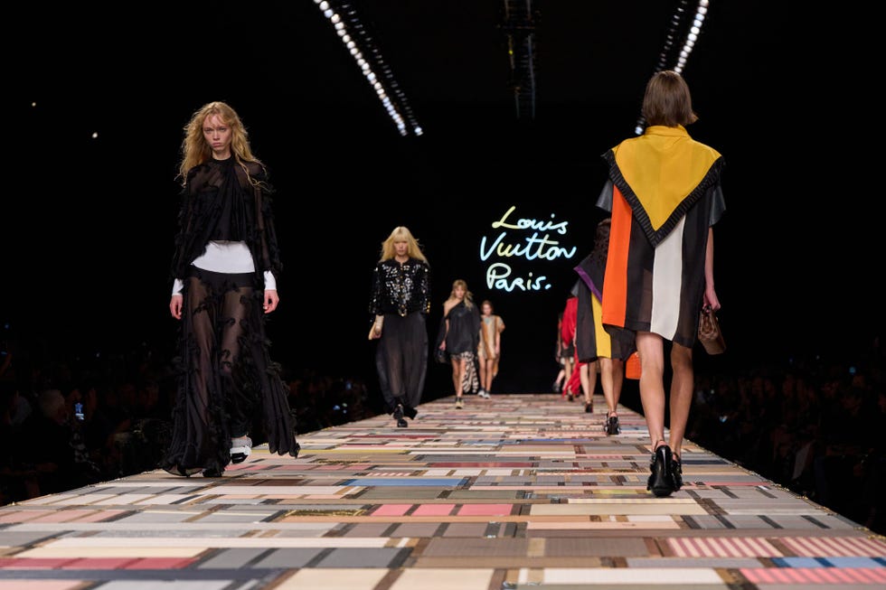 Fashion runway featuring models wearing designer outfits with a Louis Vuitton backdrop