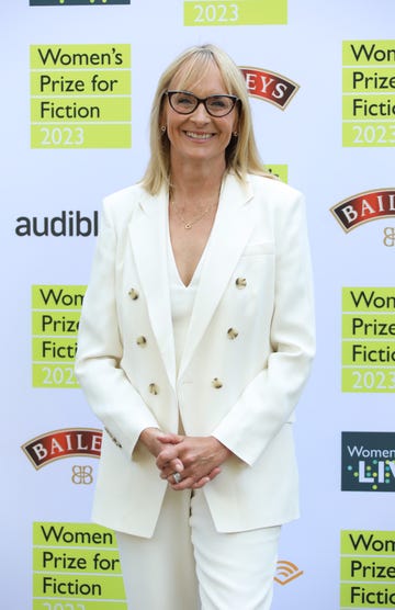 louise minchin, a woman stands clasping hands in front of her and smiles at the camera, she has shoulder length blonde hair worn down and wears a cream suit with black glasses and gold jewellery